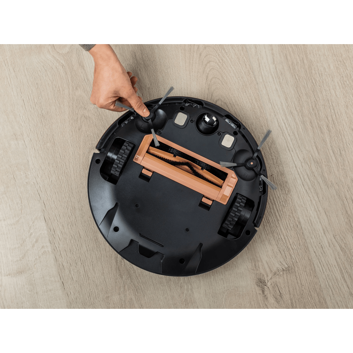 Una mano limpia la parte inferior de un robot aspirador negro en un suelo de madera.