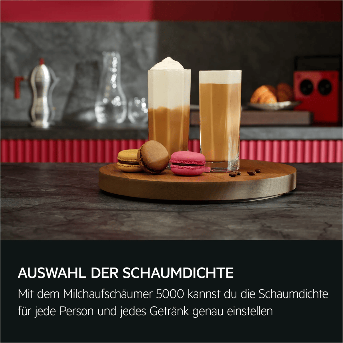 Zwei hohe Kaffeemäsche mit Schaum und Macarons auf einem Holzbrett in einer Küche.