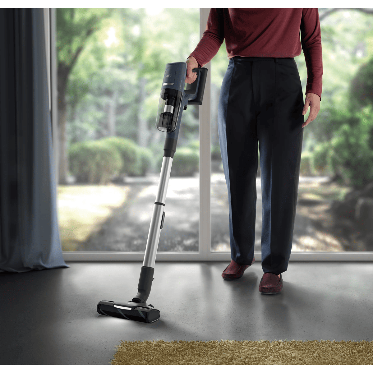 Una persona con camisa roja y pantalones oscuros aspira un suelo gris en una habitación luminosa con ventanales.