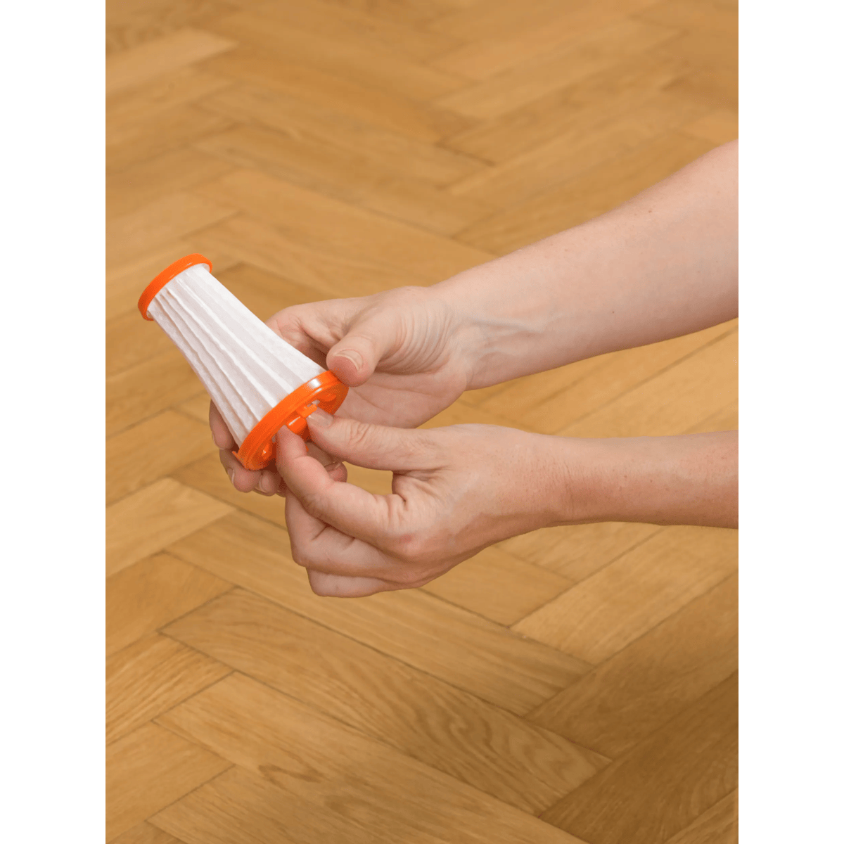 Le mani tengono un piccolo filtro per aspirapolvere bianco con bordo arancione su uno sfondo in legno.