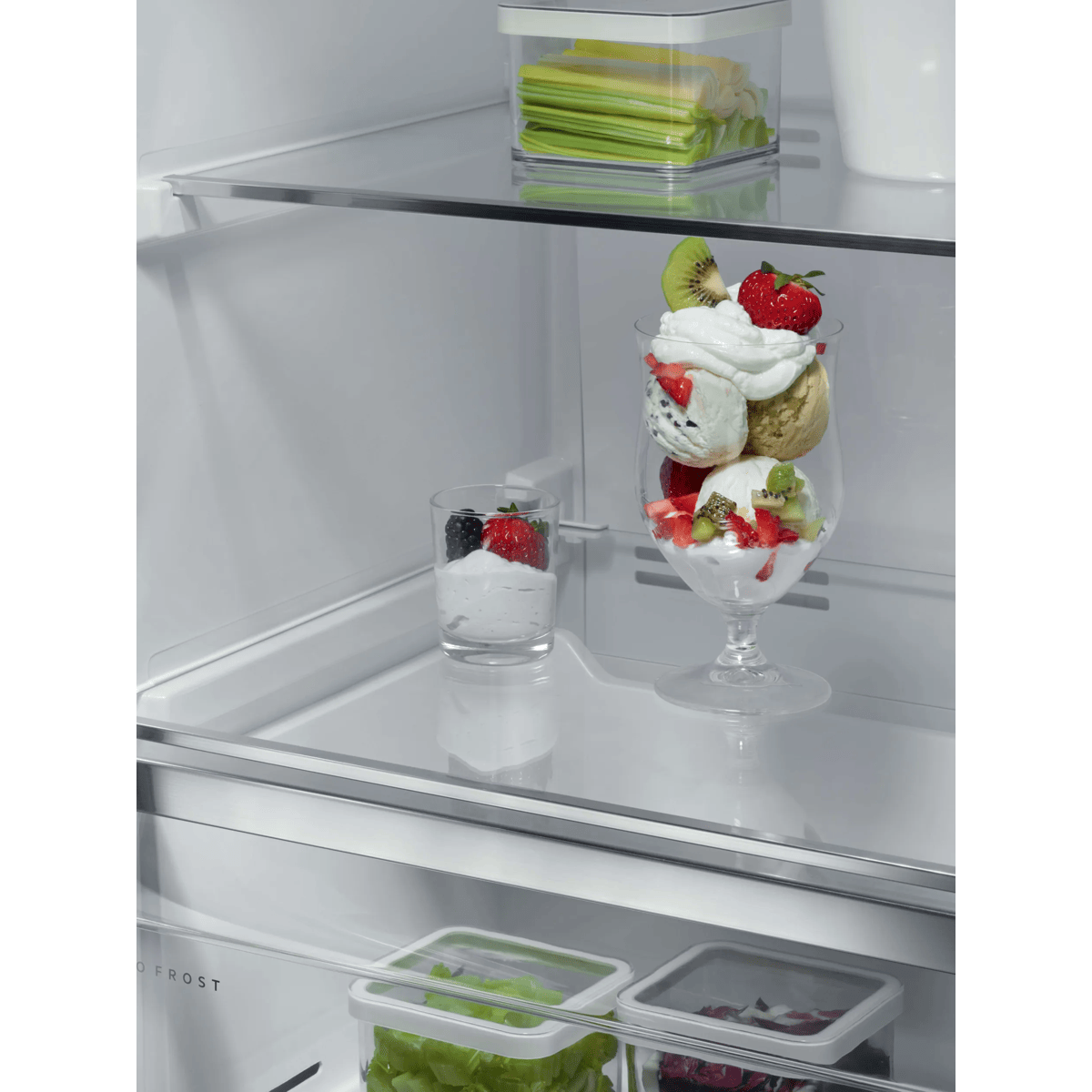 Un refrigerador blanco con estantes de vidrio contiene postres en vasos, fresas, kiwi y recipientes con verduras.