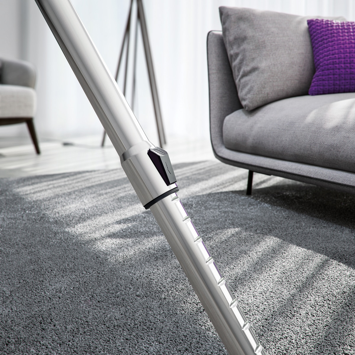 Un tubo de aspiradora plateado en primer plano, con sofá gris, cojín morado y alfombra gris al fondo.