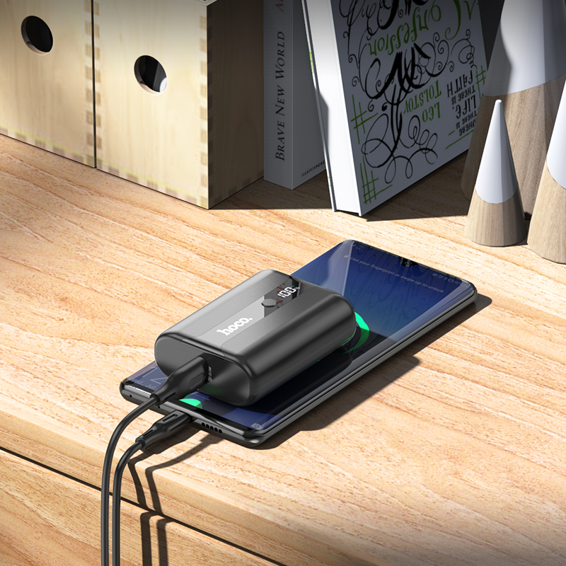 Una batería externa negra con pantalla digital carga un smartphone en un escritorio.