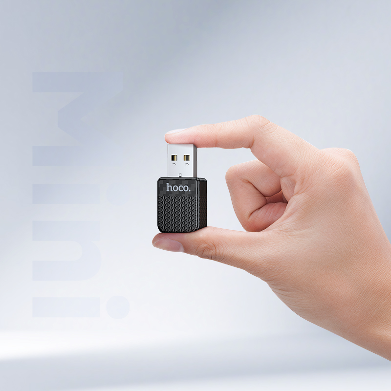 Una mano sostiene un pequeño adaptador USB hoco negro y plateado sobre un fondo gris claro.