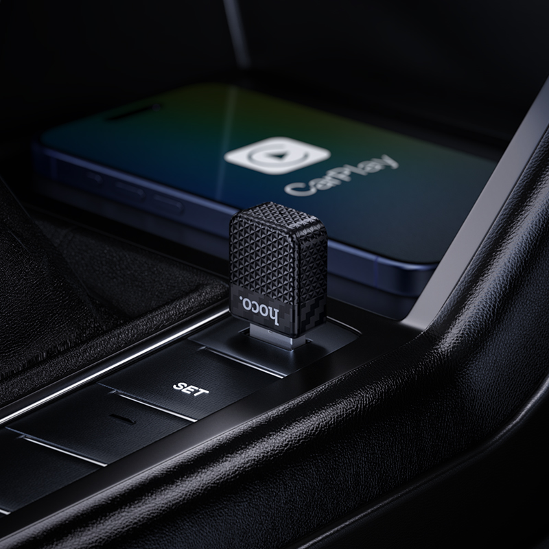 Un adaptador CarPlay inalambrico Hoco negro enchufado en la consola del coche, con un telefono inteligente al fondo.