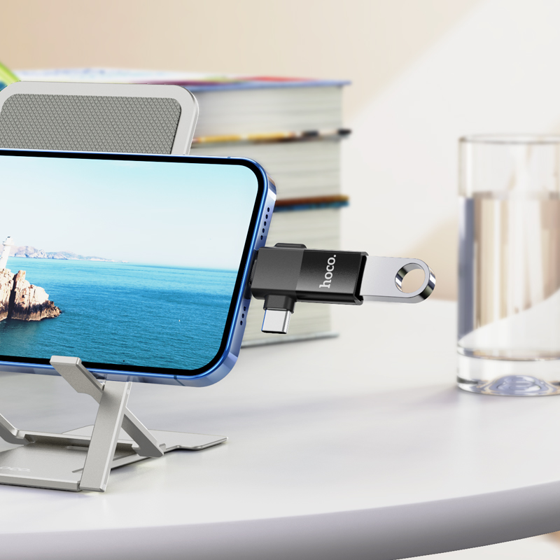 Un adaptador USB Hoco negro enchufado a un smartphone en un soporte de metal, con libros y un vaso de agua al fondo.
