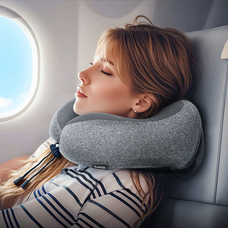 Mujer durmiendo en un asiento de avión con un cojín cervical gris junto a la ventana.