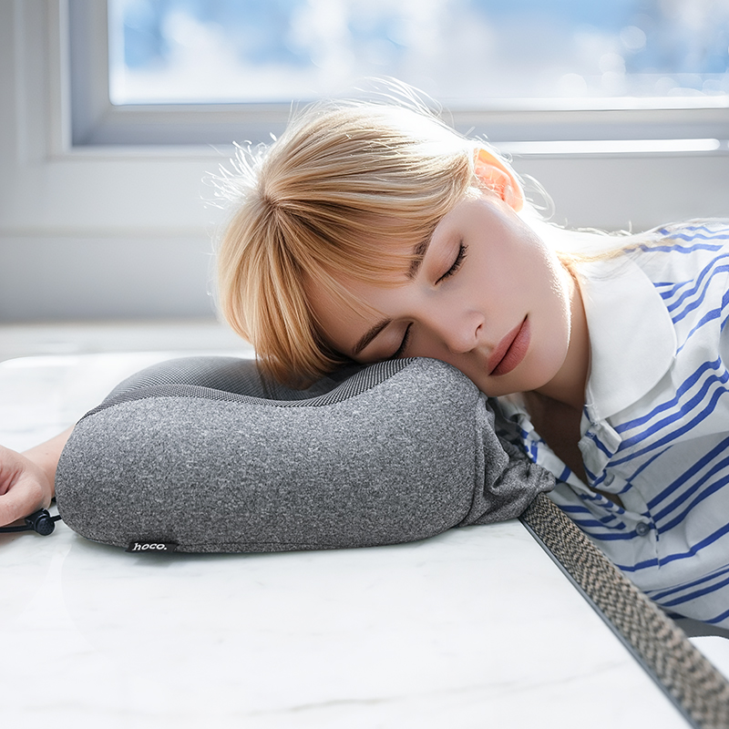 Una mujer rubia duerme plácidamente apoyando la cabeza en una almohada de viaje gris sobre una mesa blanca.