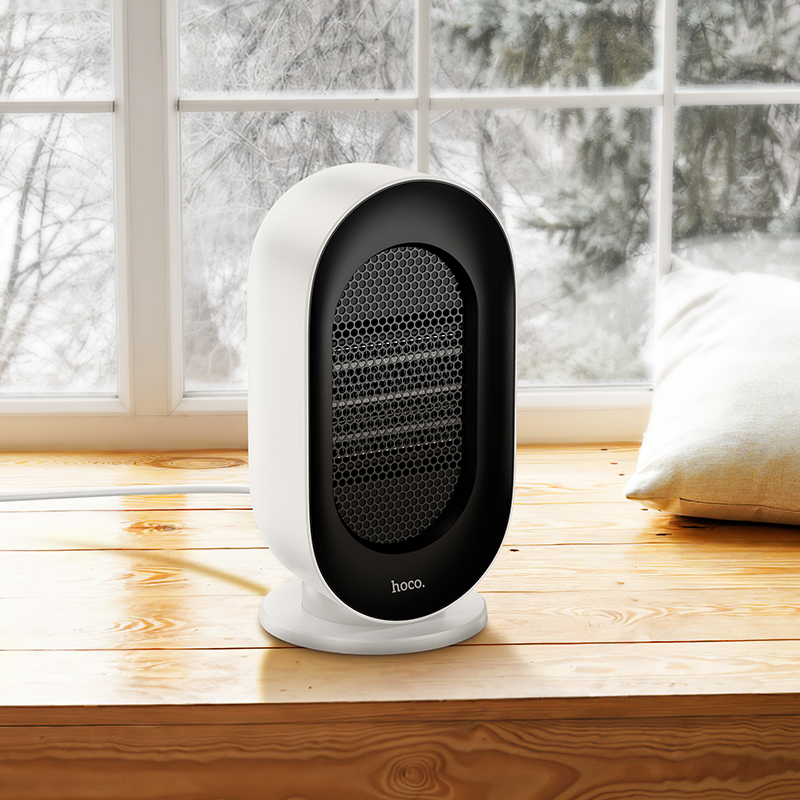 Un calefactor eléctrico hoco blanco y negro sobre un alféizar de madera ante una ventana nevada con un cojín.