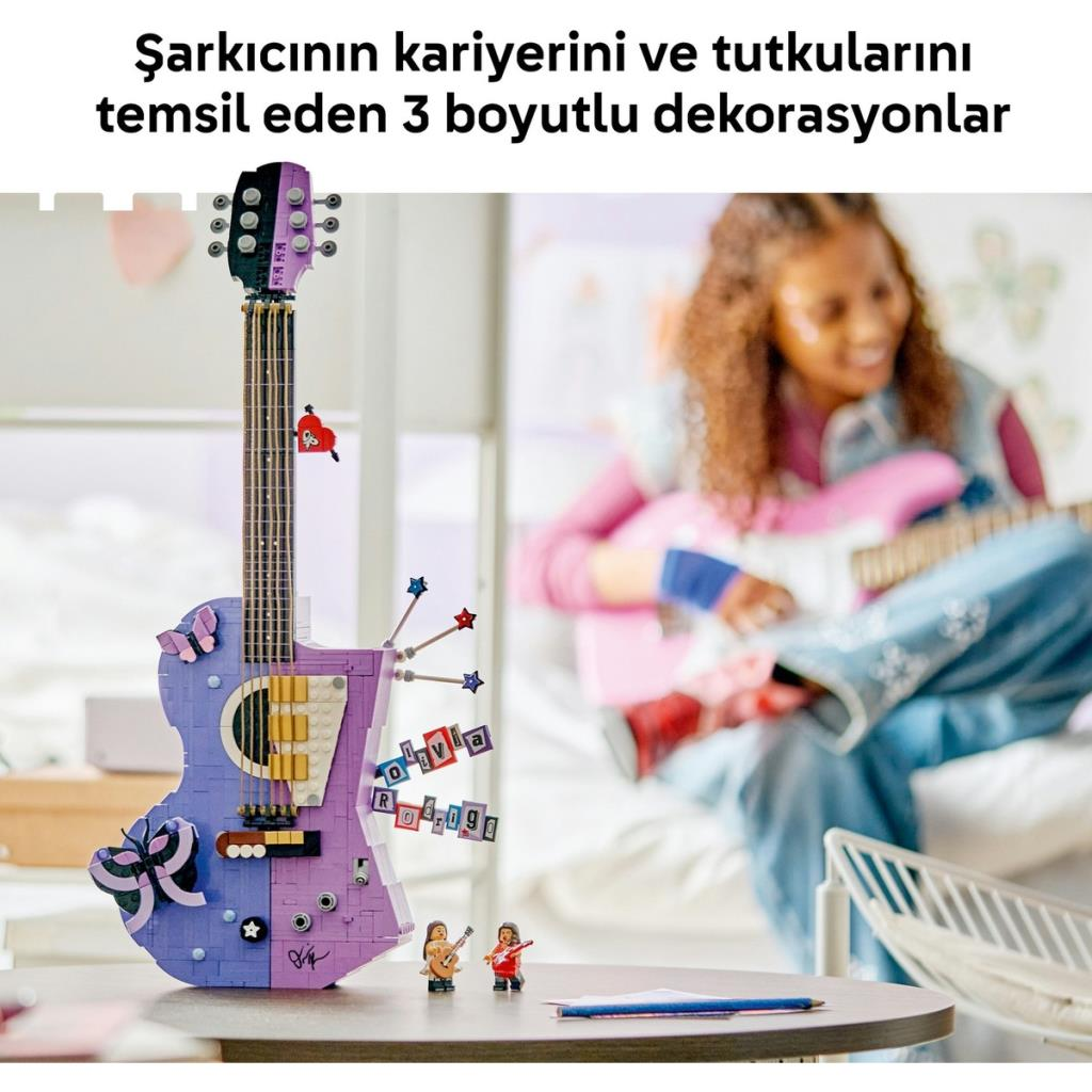 Bir masada mor renkli LEGO gitar duruyor, arka planda ise başka bir gitar tutan genç bir kişi var.