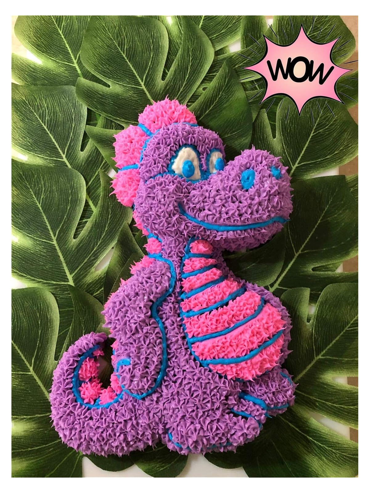 Eine lila-pinke Dinosaurier-Torte mit blauen Akzenten, platziert auf grünen tropischen Blättern.