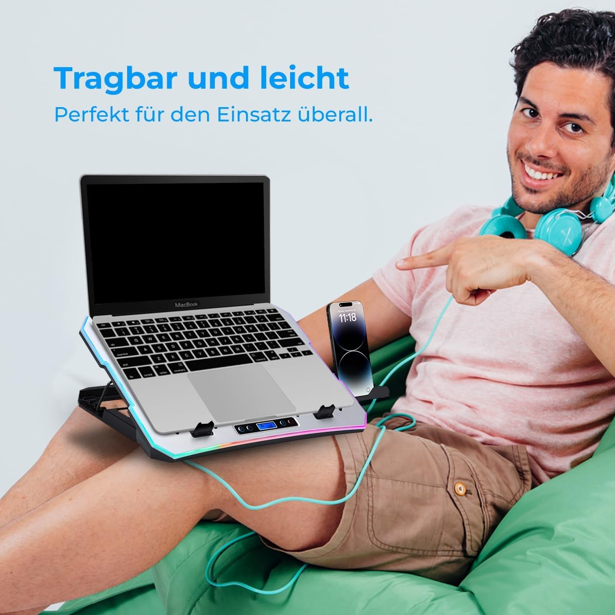Mann sitzt auf einem grünen Kissen, zeigt auf seinen Laptop auf einem beleuchteten Ständer, mit Handy daneben.