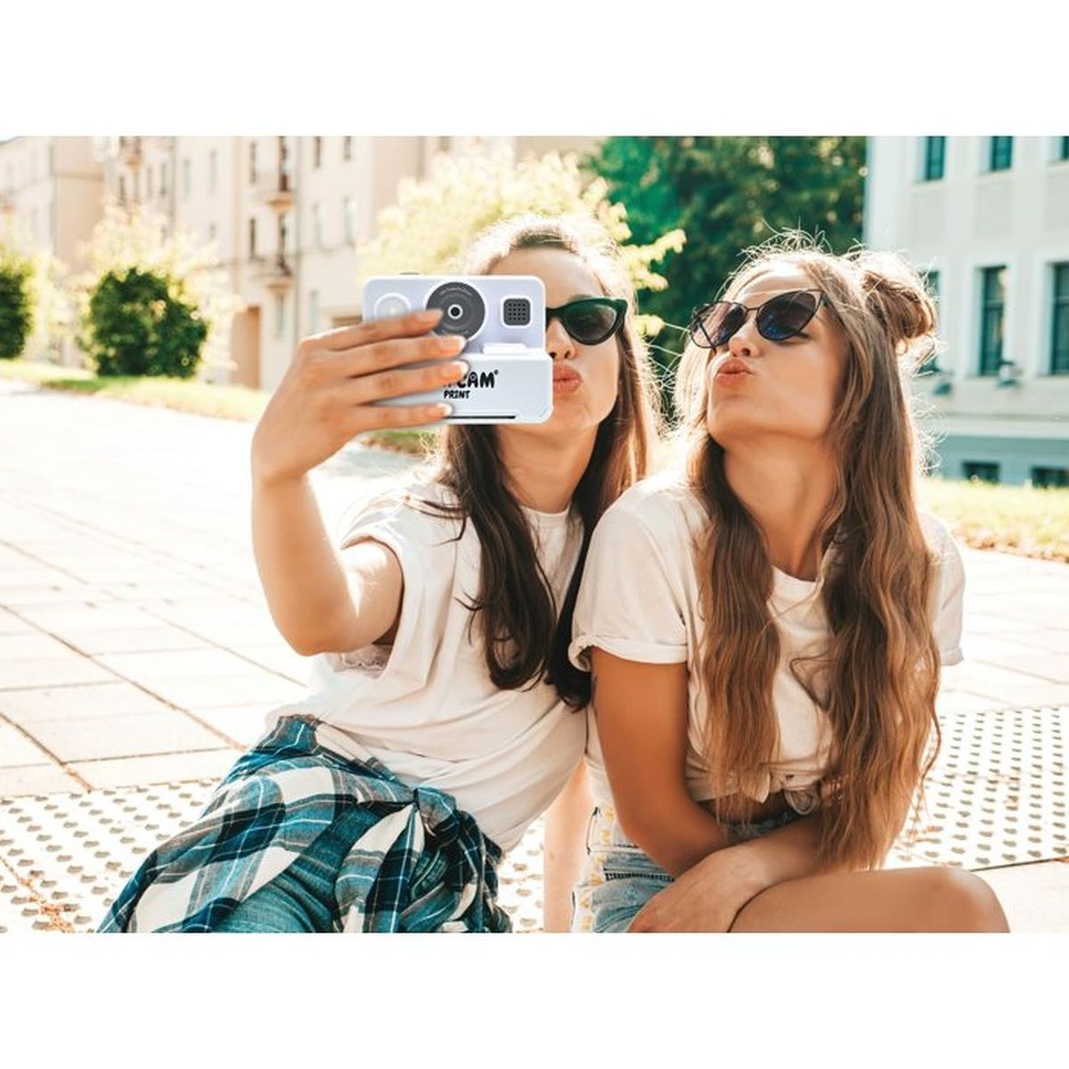 Zwei junge Frauen in Sonnenbrillen machen draußen ein Selfie mit einer kleinen Kamera und spitzen den Mund.