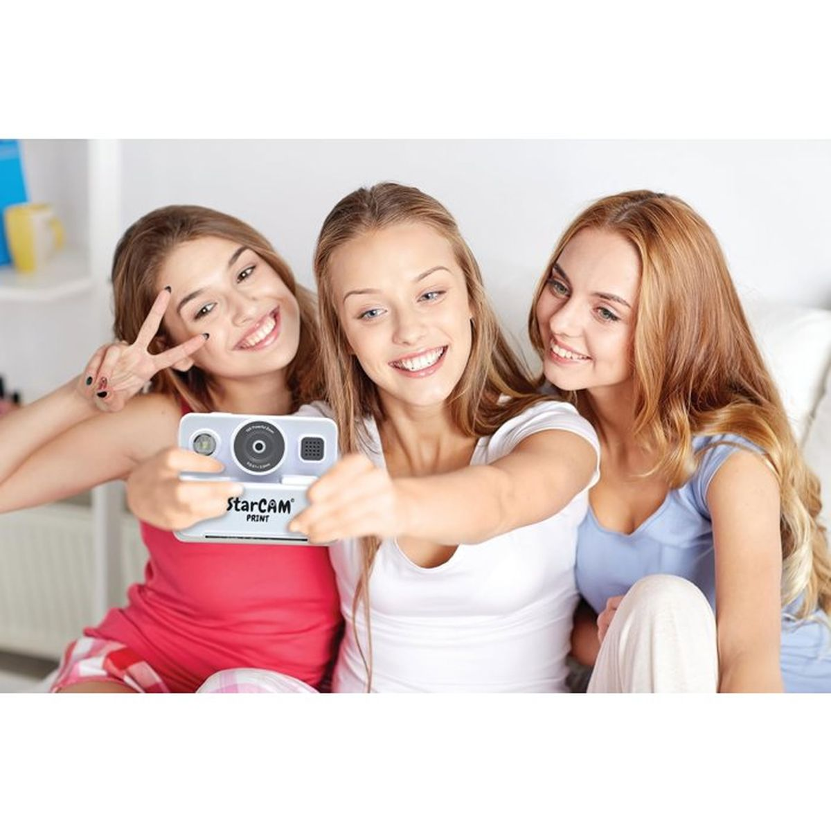 Drei lächelnde junge Frauen machen drinnen ein Selfie mit einer kleinen weißen Sofortbildkamera.