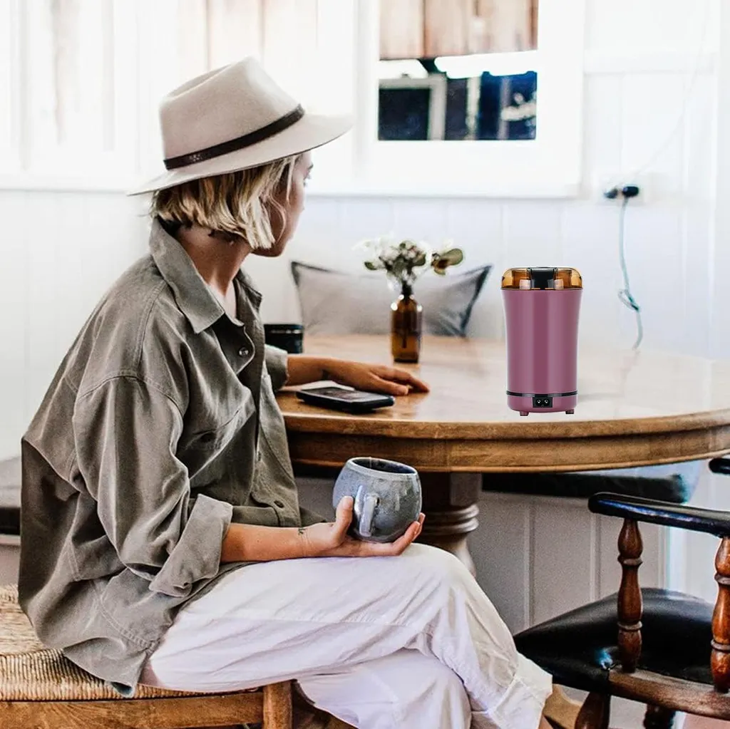 Eine Frau mit Hut sitzt an einem Holztisch mit einer Tasse, daneben eine Kaffeemühle.
