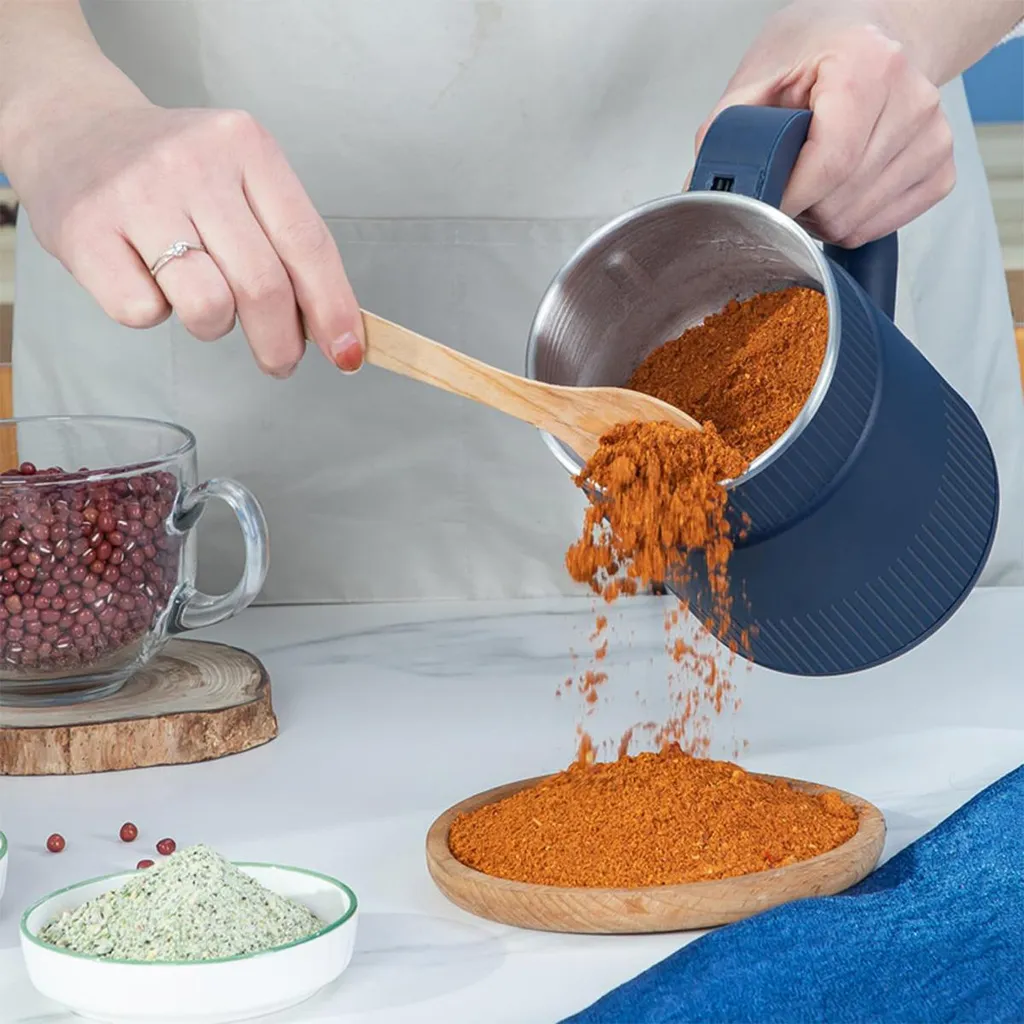 Eine Person gießt gemahlenes rotes Gewürz aus einem blauen Behälter auf einen Holzteller, daneben rote Bohnen.