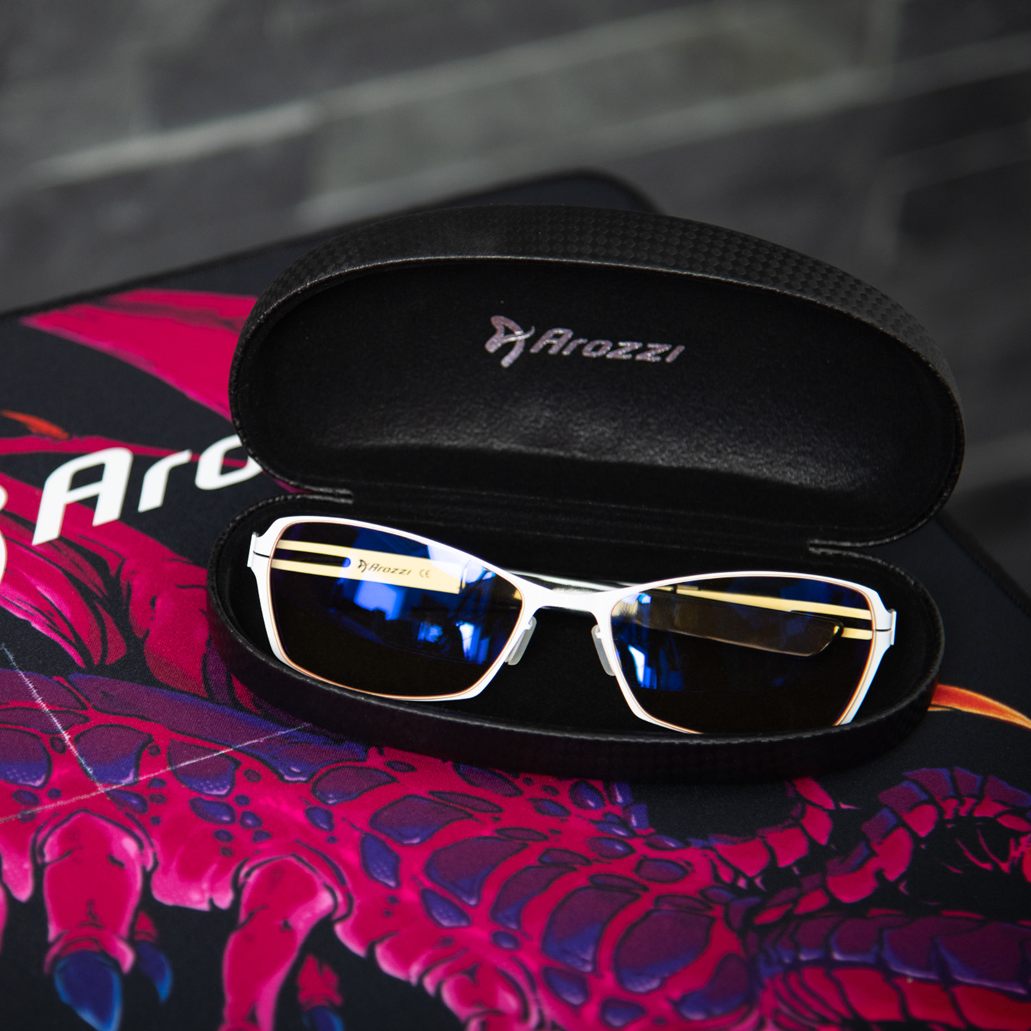 Occhiali da gaming bianchi con lenti blu in astuccio Arozzi nero aperto su tappetino rosso e nero.