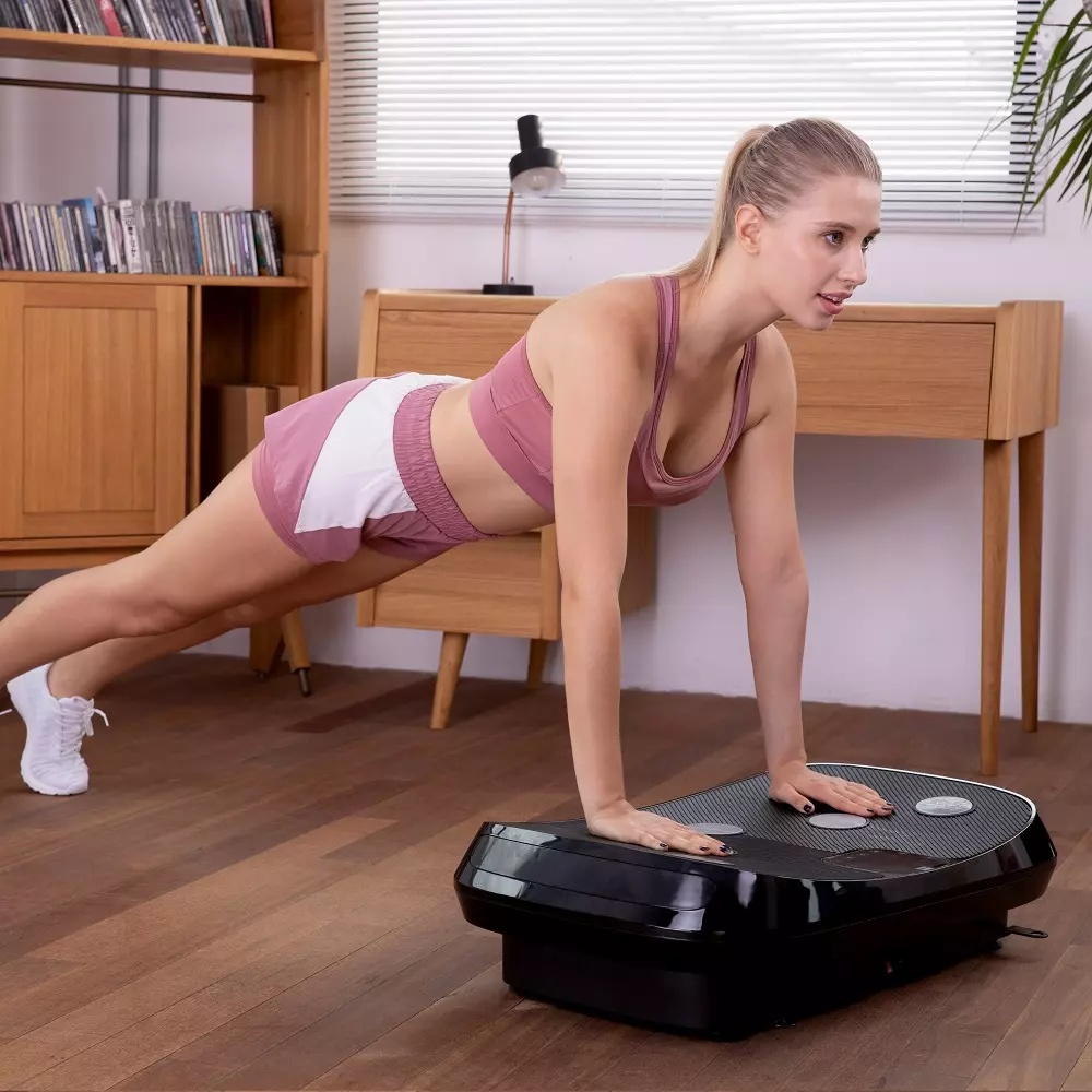 Eine Blondine macht Liegestütze auf einer Vibrationsplatte in einem hellen Raum.