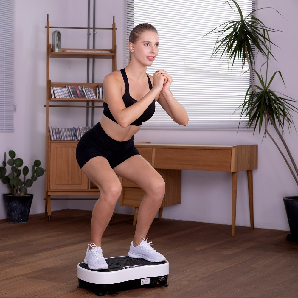 Eine Frau in Sportkleidung macht Kniebeugen auf einer weißen Vibrationsplatte in einem hellen Raum.