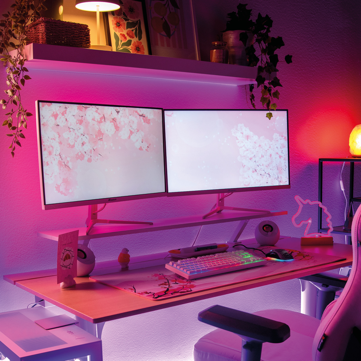 Ein Schreibtisch mit zwei Monitoren, einer rosa Kirschblüten-Tastatur, RGB-Beleuchtung und Pflanzen im Hintergrund.