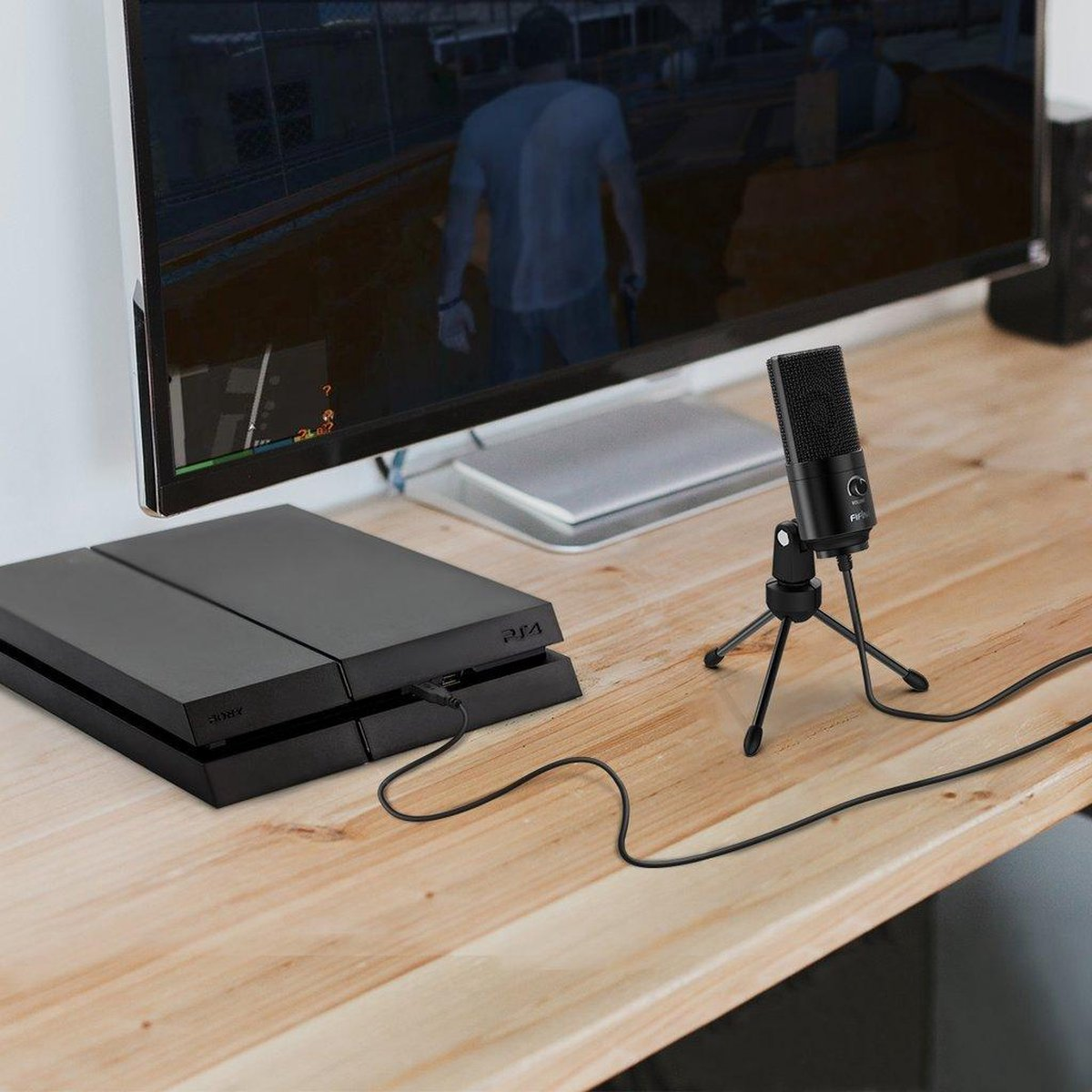 Een zwarte microfoon op een klein statief en een PS4 op een houten bureau voor een groot scherm.