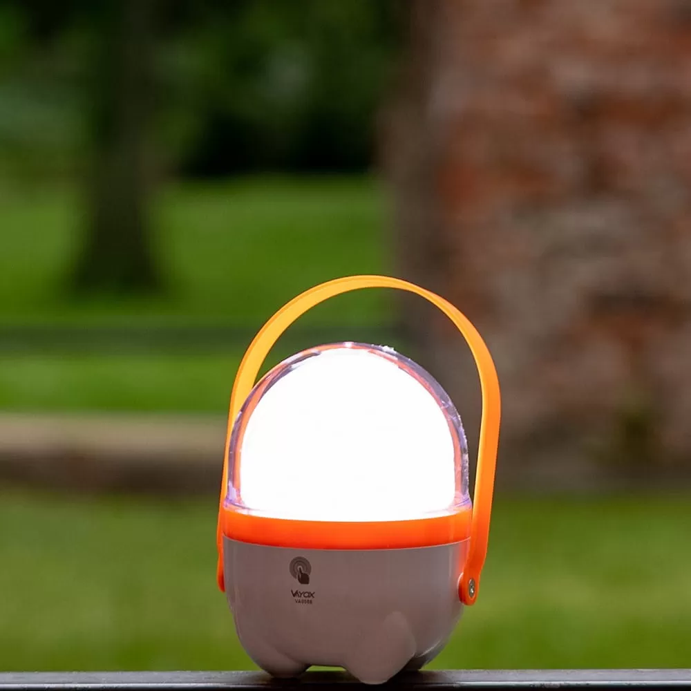 Mała, przenośna, biało-pomarańczowa lampka LED z uchwytem stoi na stoliku na świeżym powietrzu.