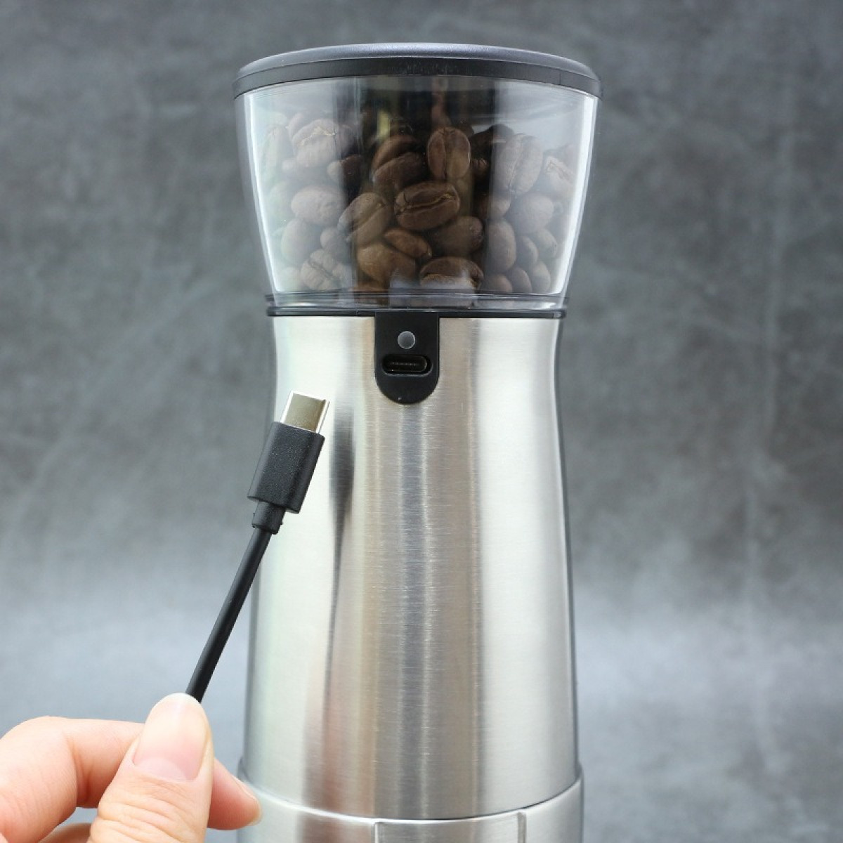 Eine Hand hält ein USB-Kabel vor eine elektrische Kaffeemühle mit Bohnen.