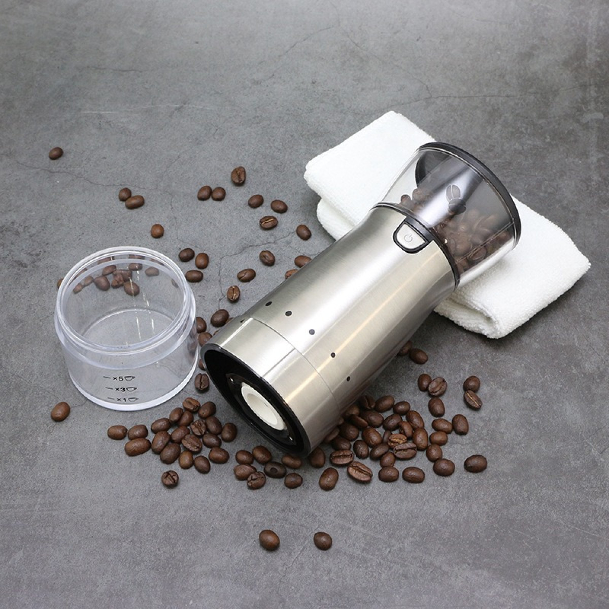 Eine Kaffeemühle aus Edelstahl liegt auf einer grauen Oberfläche mit Kaffeebohnen und einem weißen Tuch.
