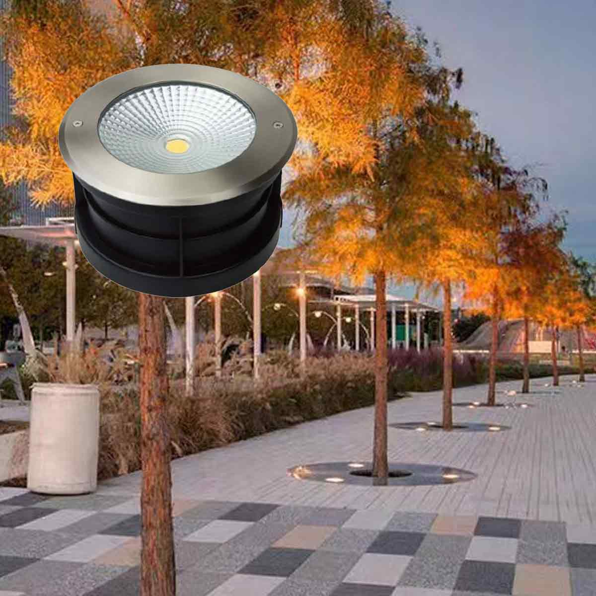 Runde Einbauleuchte im Vordergrund, beleuchteter Parkpfad mit Bäumen und Lichtern im Hintergrund bei Dämmerung.