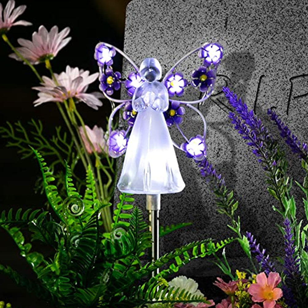 Beleuchtete Engelsfigur mit violetten Blumenflügeln vor einem Grabstein.