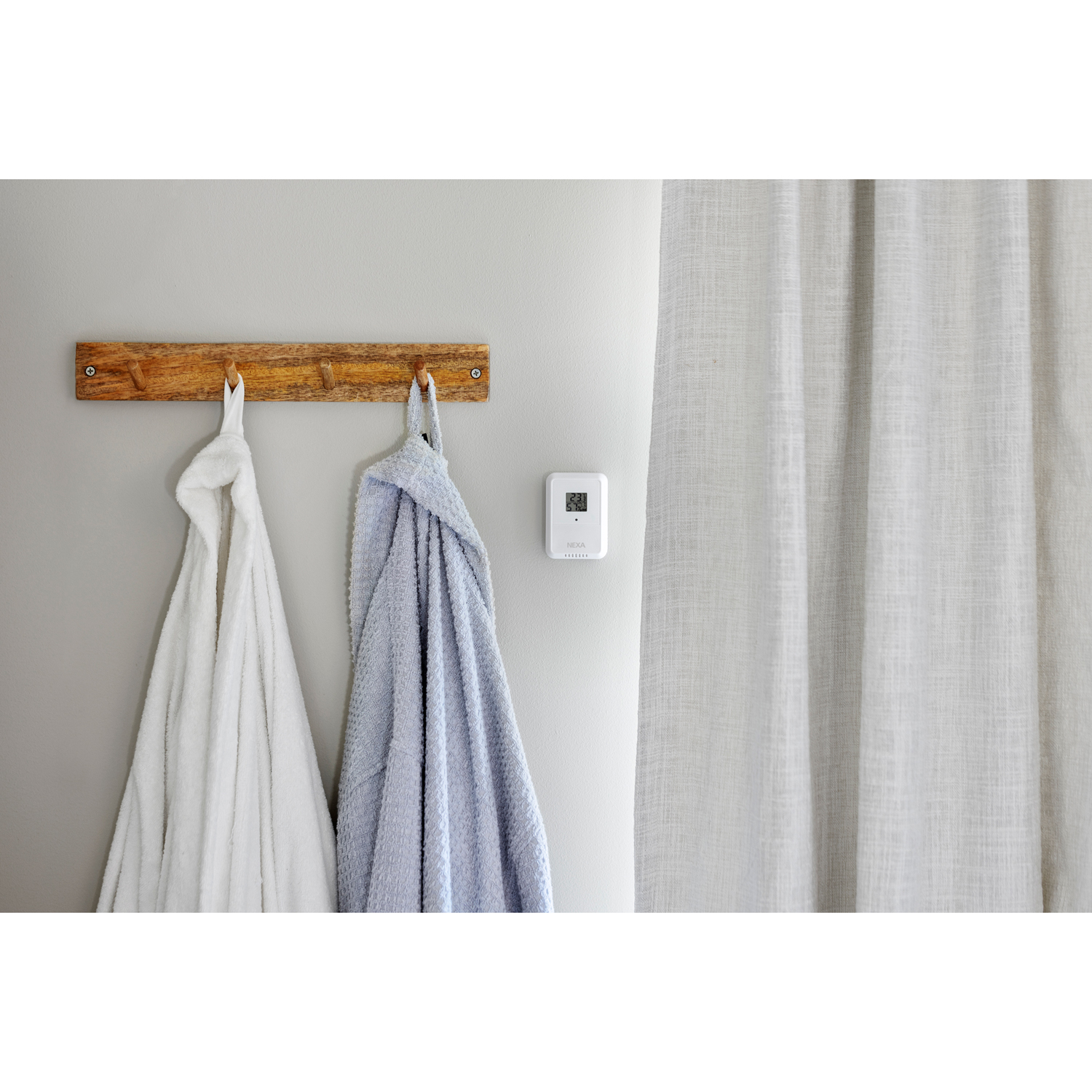 Ein weißer und ein blauer Bademantel hängen an einem Holzhalter an der Wand neben einem Thermometer und einem Vorhang.