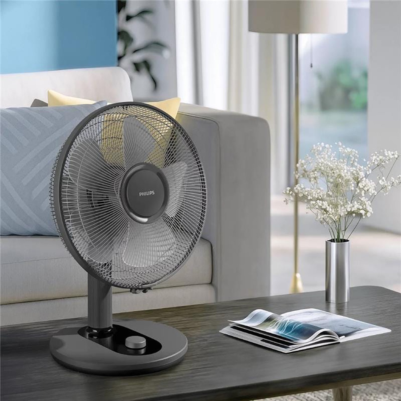 Ein grauer Philips Tischventilator steht auf einem Tisch vor einem Sofa mit Kissen in einem modernen Wohnzimmer.