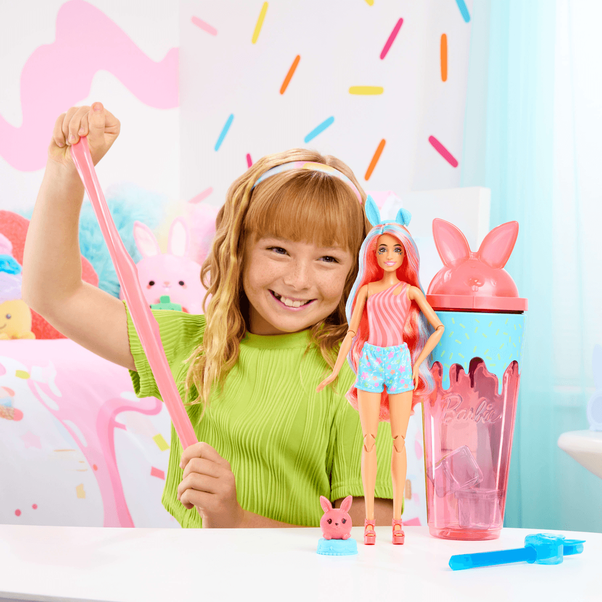 Ein lächelndes Mädchen im grünen Shirt dehnt rosa Schleim neben einer Barbie-Puppe und einem Hasenbecher.