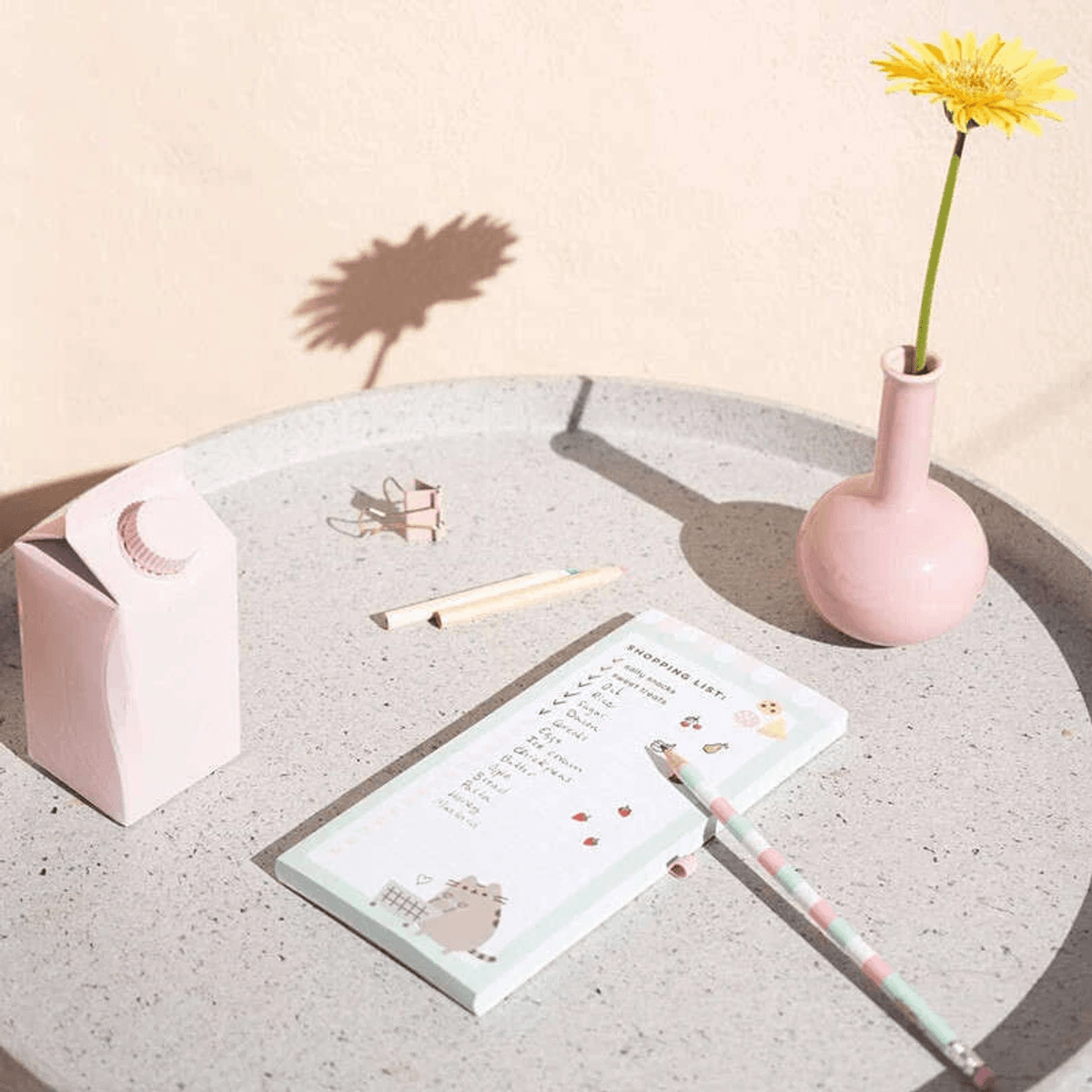 Ein rosafarbener Einkaufszettelblock, eine Blume in einer rosa Vase und ein rosa Milchkarton auf einem grauen Tablett.