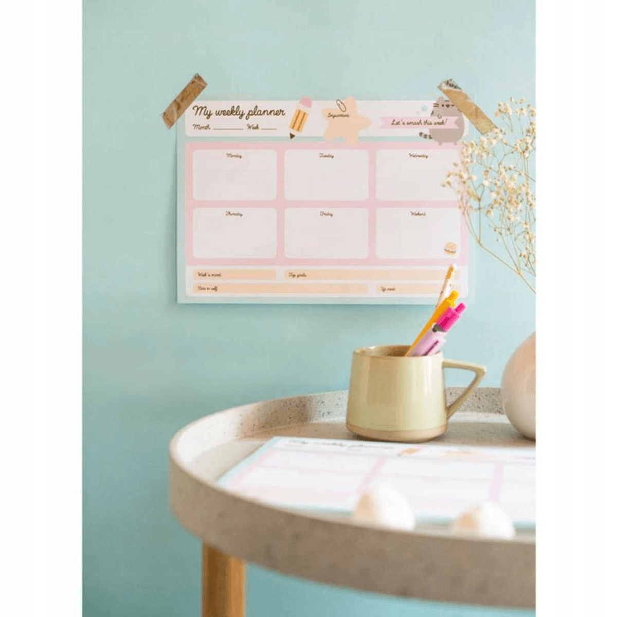 Ein pastelnder Wochenplaner an der mintgrünen Wand, ein Becher mit Stiften und Blumen auf einem Tisch.