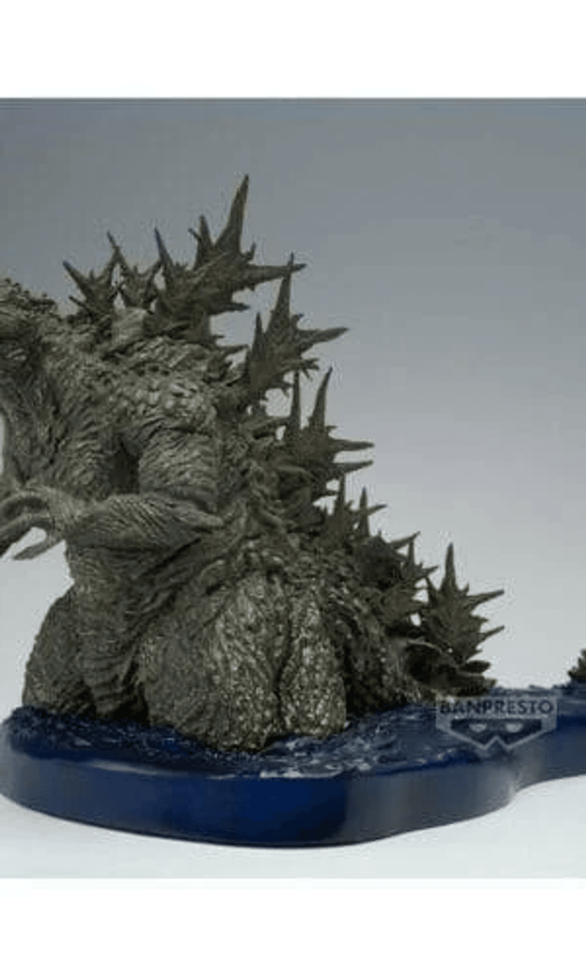 Eine graue Godzilla-Figur steht auf einem blauen Sockel vor grauem Hintergrund.
