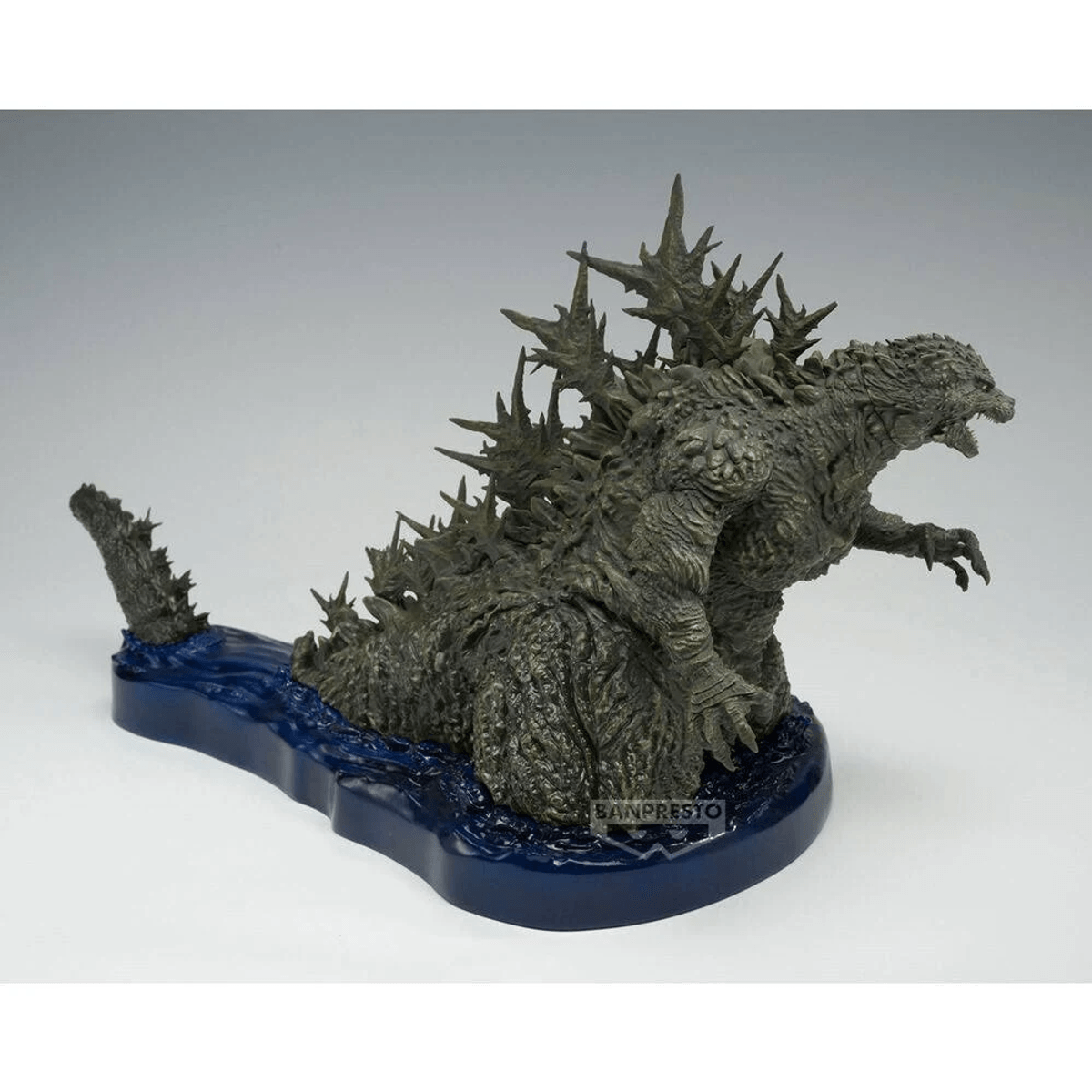 Eine graue Godzilla-Figur steht auf einem blauen, wassersockelartigen Ständer vor weißem Hintergrund.