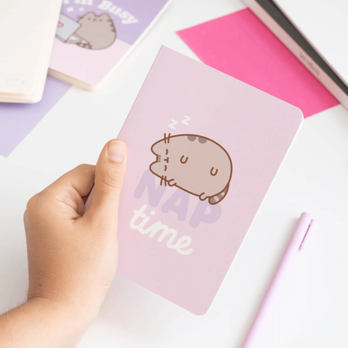 Eine Hand hält ein rosafarbenes Notizbuch mit einer schlafenden Katze und der Aufschrift NAP time, auf weißem Tisch.