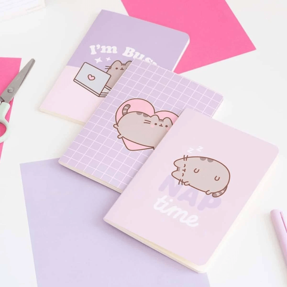 Drei pastellviolette Notizbücher mit Katzenmotiven, umgeben von rosa Papier und Schere.