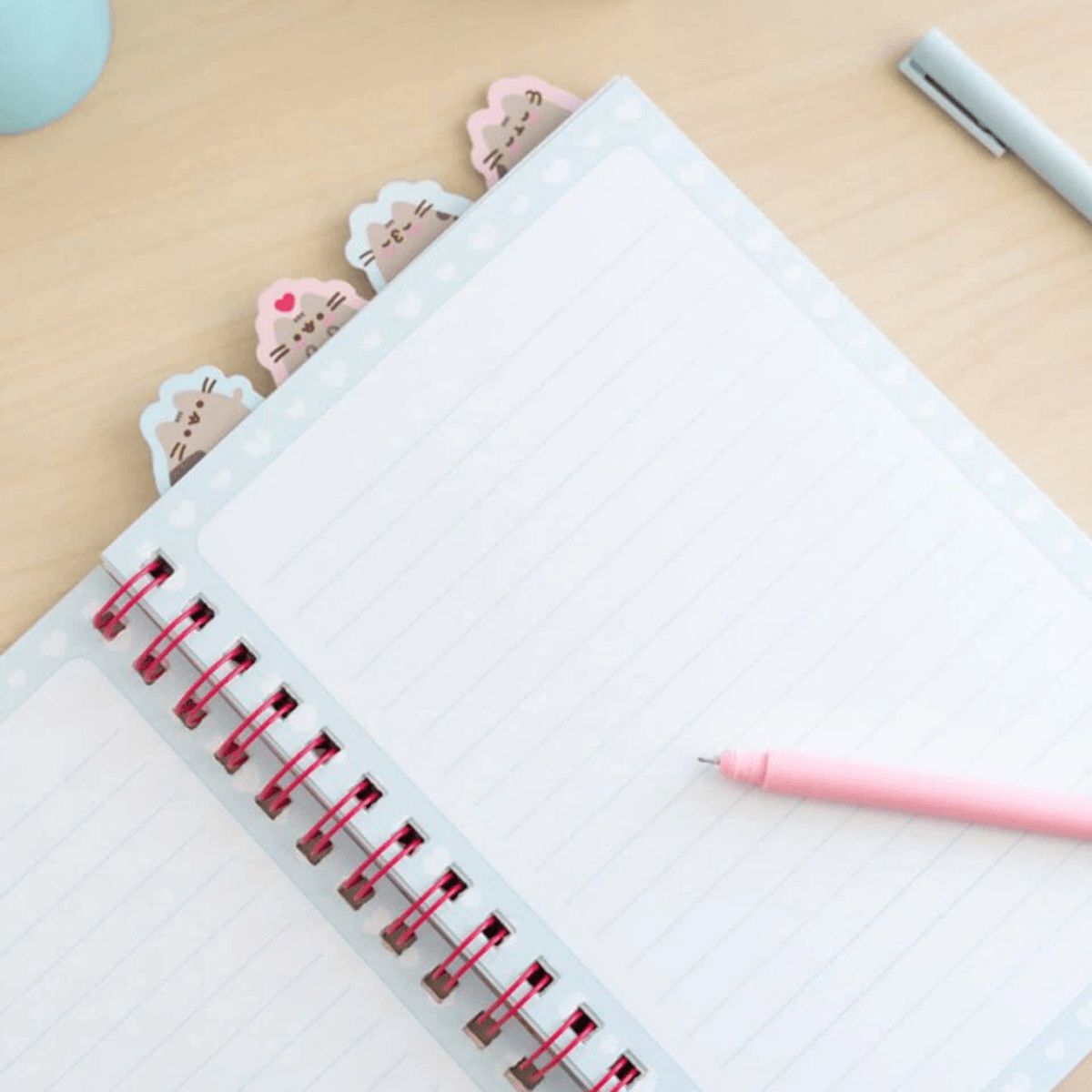 Ein geöffnetes liniertes Notizbuch mit katzenförmigen Klebezetteln und einem rosa Stift auf einem Holzschreibtisch.