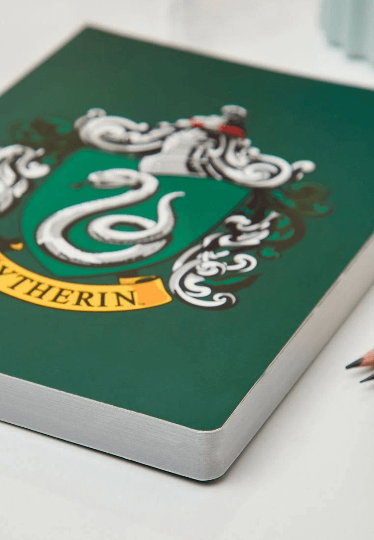 Ein grünes Slytherin-Notizbuch mit silbernem Schlangenwappen auf weißem Grund.