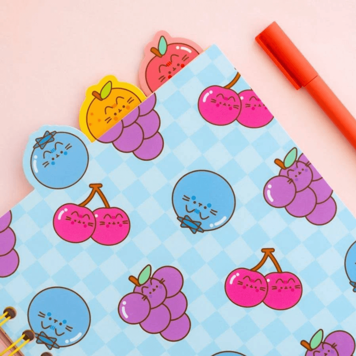 Ein blaues kariertes Notizbuch mit niedlichen Frucht-Motiven und einem roten Stift auf rosa Hintergrund.