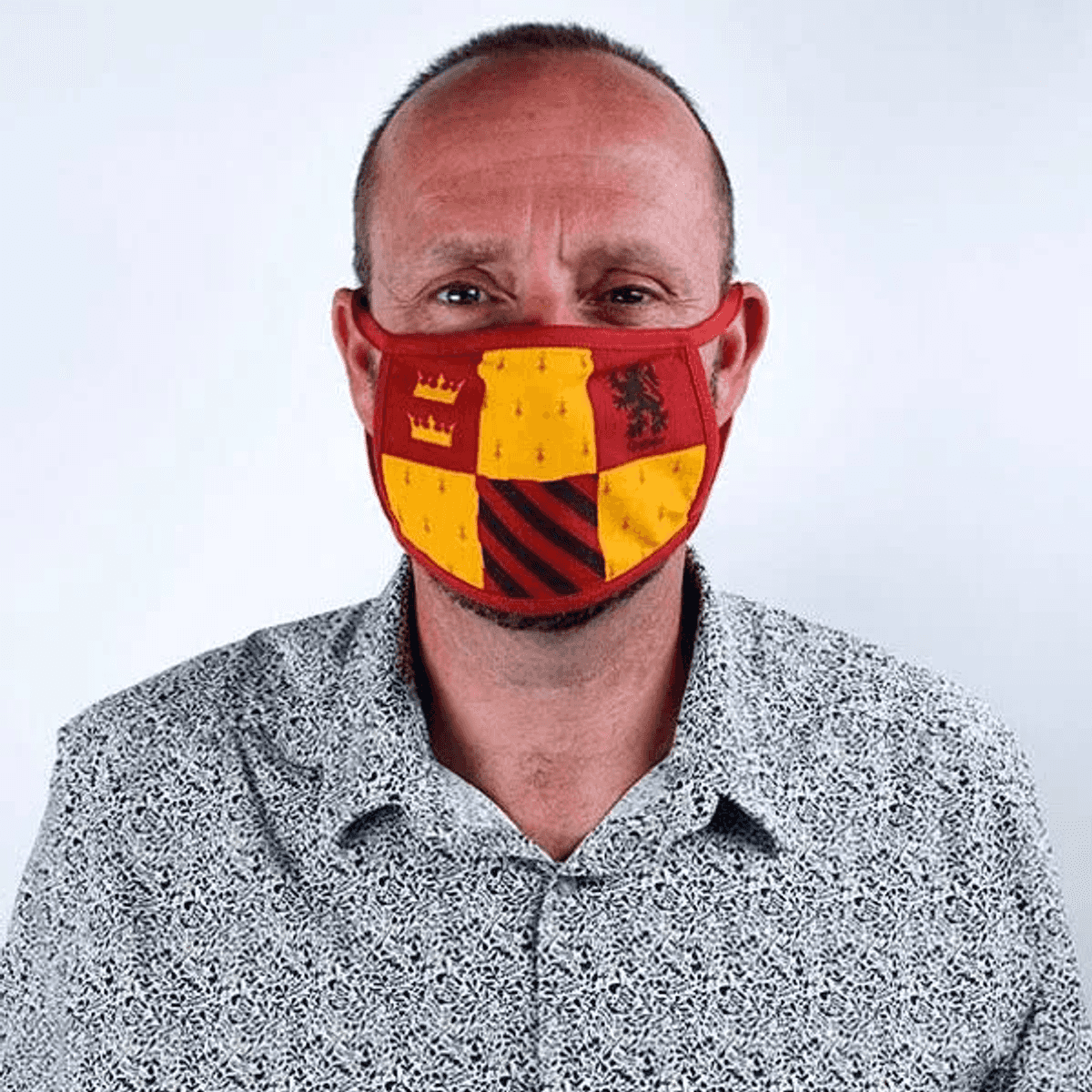 Ein Mann mit einer Gesichtsmaske mit rotem und gelbem Wappenemblem und einem gemusterten Hemd.