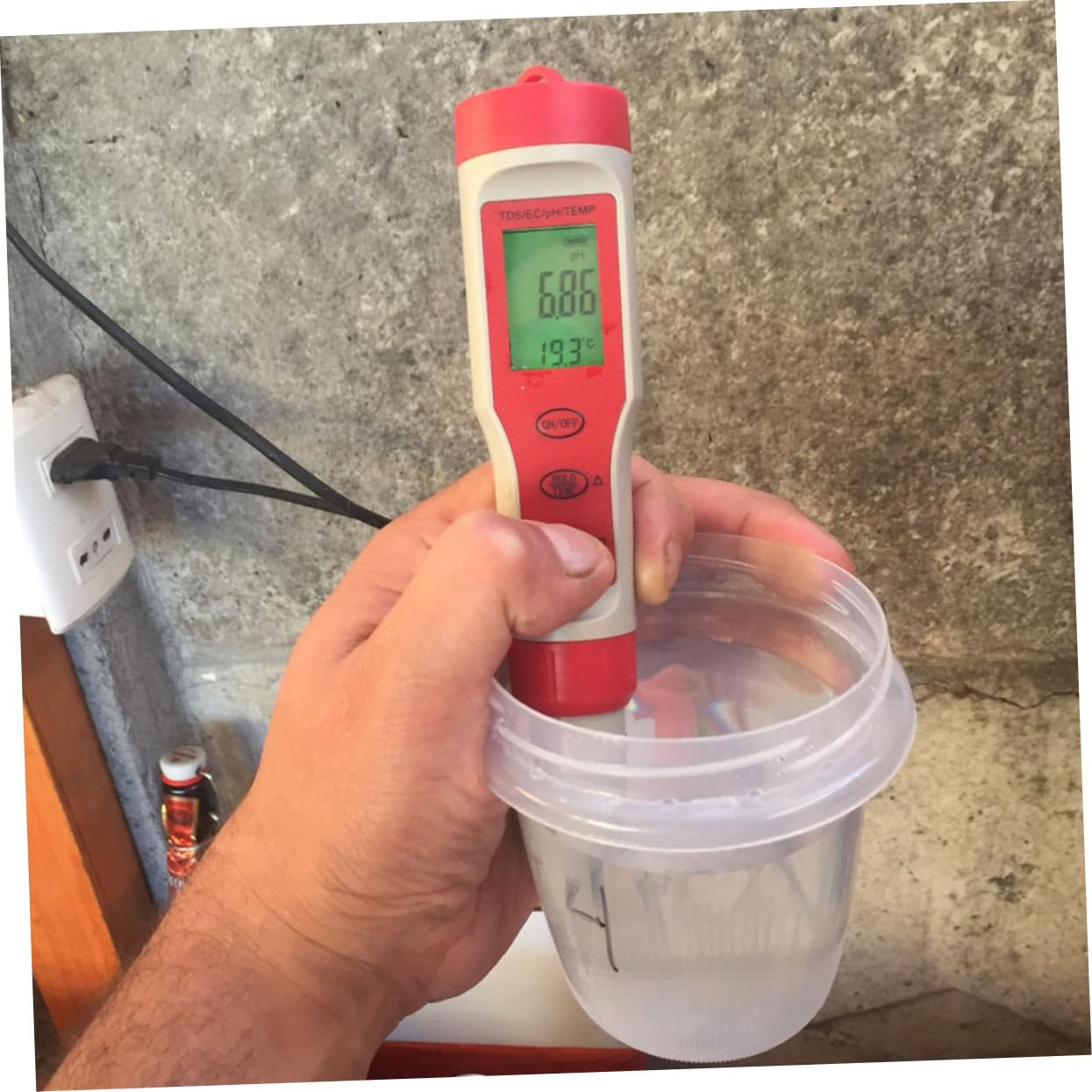 Eine Hand hält ein digitales Wassertestgerät in einem Plastikbecher vor einer Betonwand.