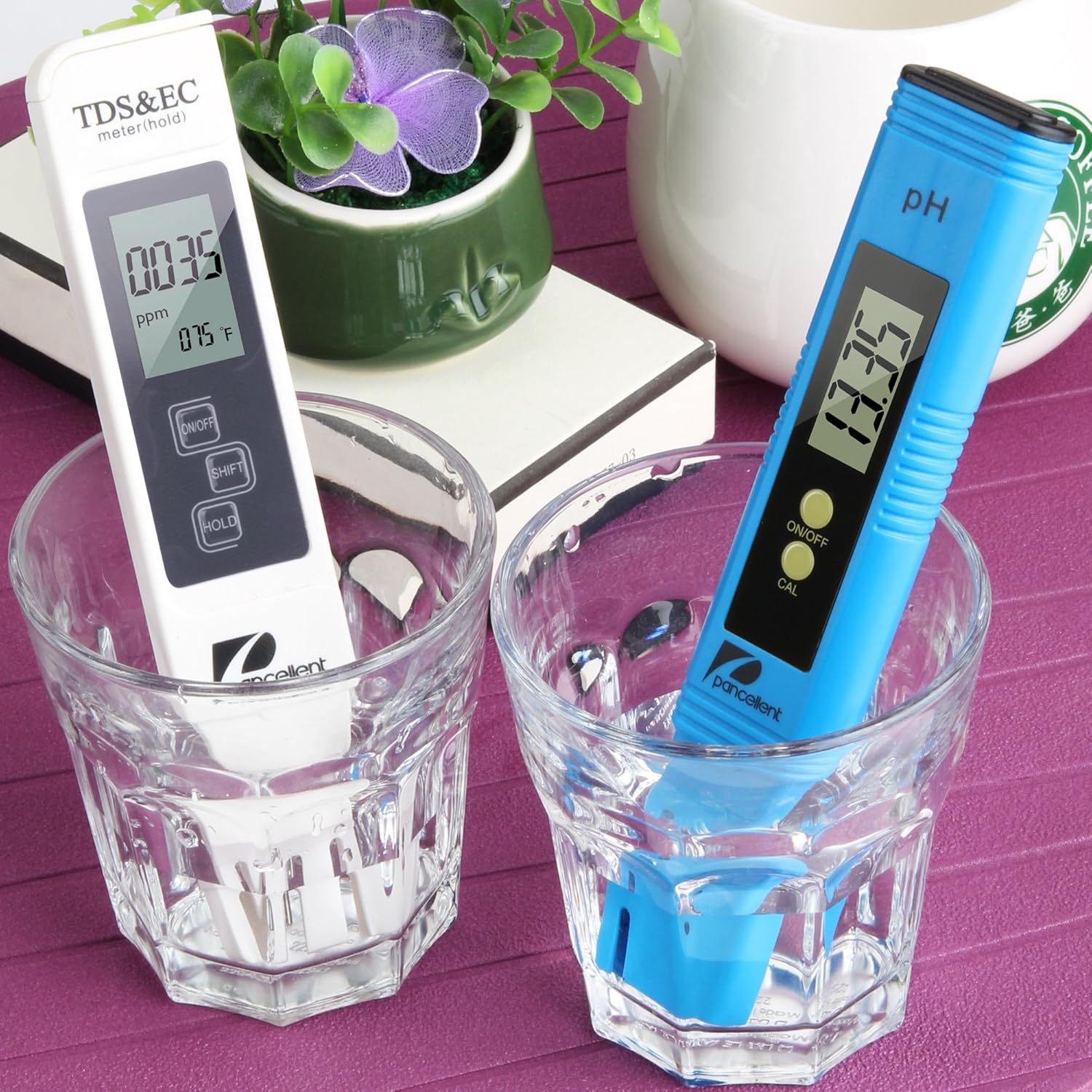 Ein weißes TDS-Meter und ein blaues pH-Meter stehen in Wassertrinkgläsern auf einem violetten Tisch.