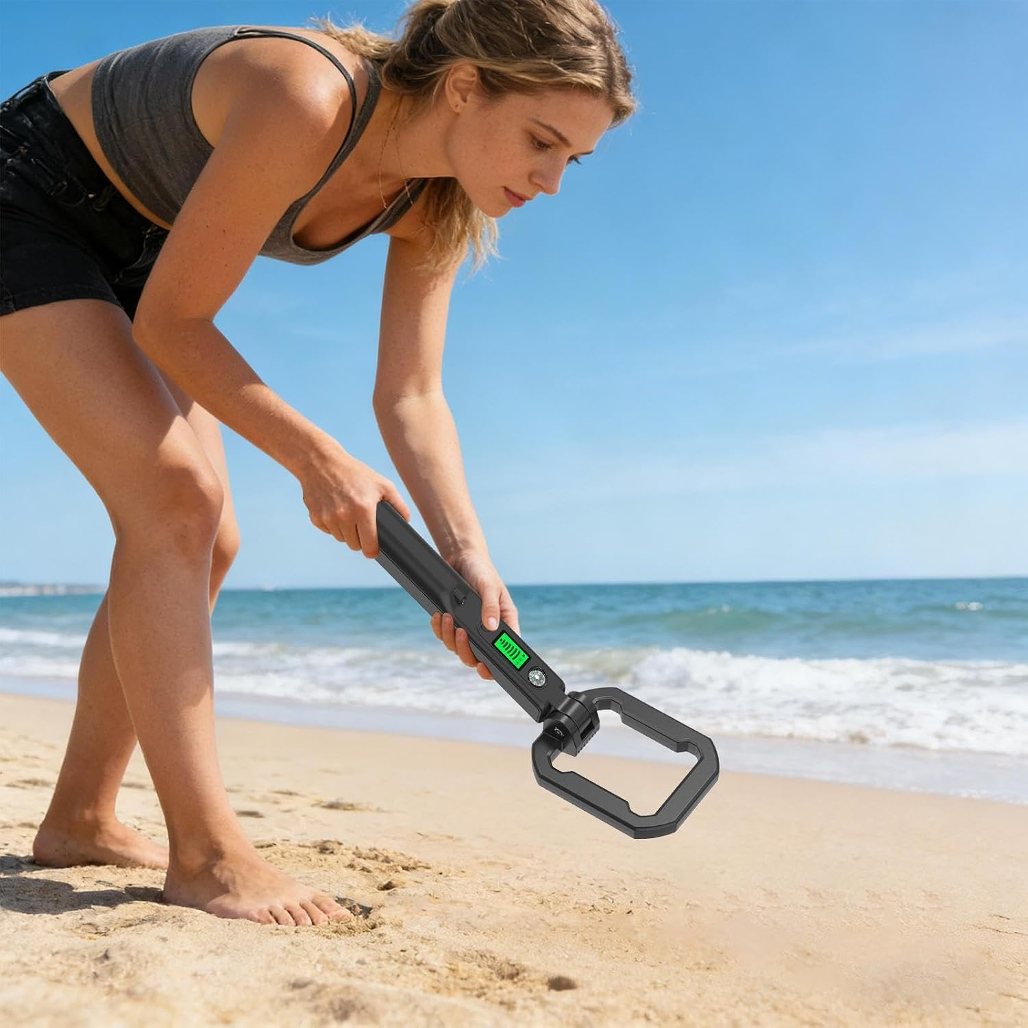 Eine Frau im grauen Oberteil hält ein kleines schwarzes Hand-Metalldetektor-Gerät am sandigen Strand vor dem Meer.