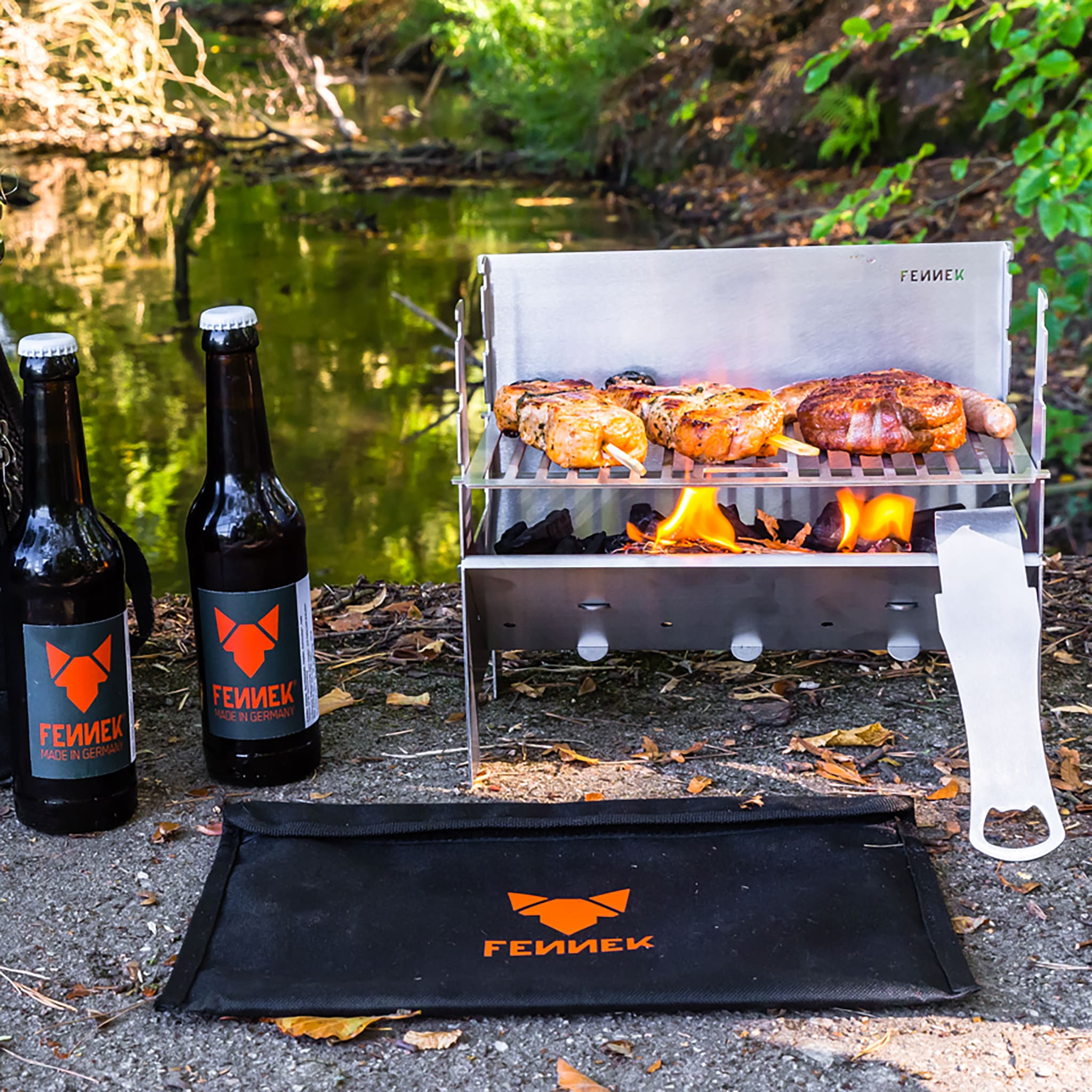 Kleiner Grill mit Fleisch, zwei Bierflaschen und eine Tragetasche am Flussufer.
