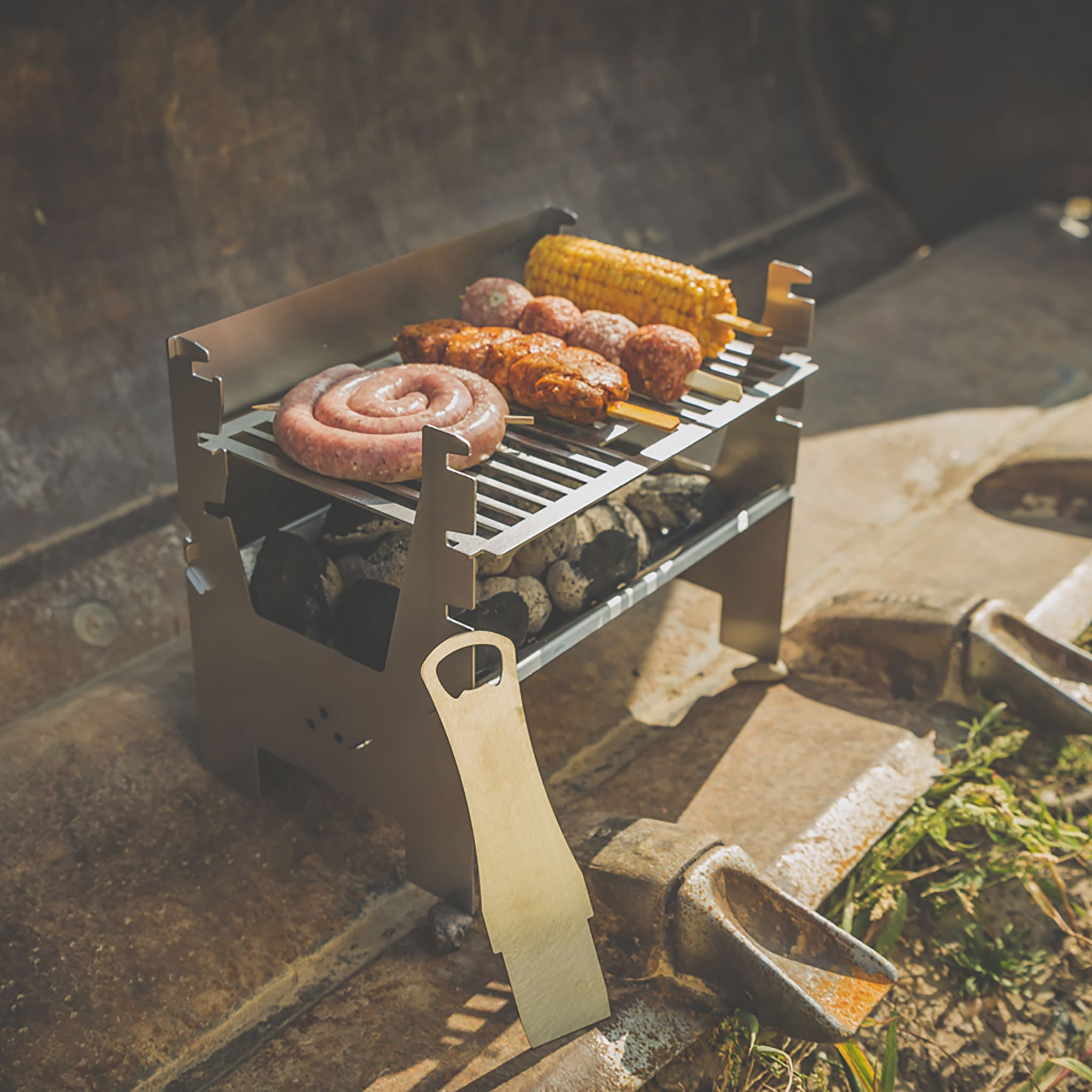 Ein kleiner Metallgrill mit einer Spirale aus Wurst, Fleischspießen und Maiskolben über heißen Kohlen.