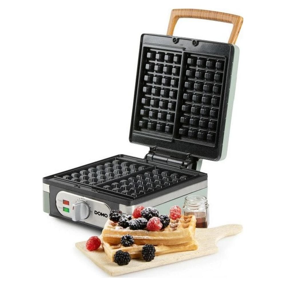 Una piastra per waffle aperta con manico in legno si trova accanto a waffle appena sfornati con bacche su un tagliere.