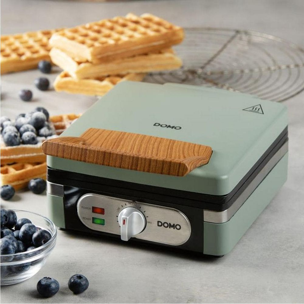 Una piastra per waffle Domo verde chiaro su un bancone grigio, circondata da waffle freschi, mirtilli e una ciotola.